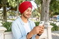 Happy and smiling indian guy in casual shirt and red national turban using smartphone outdoors Royalty Free Stock Photo