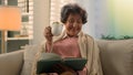 Happy smiling inspired Caucasian old woman covered with blanket on sofa drinking tea coffee with cup reading book smile