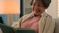 Happy smiling laughing Caucasian old woman reading bestseller book peace at home covered with blanket senior mature lady Royalty Free Stock Photo