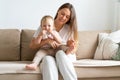 Smiling light-haired mother sitting with blond blue-eyed toddler eating socks on sofa at home Royalty Free Stock Photo