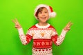 Happy smiling little boy in Santa Claus hat, Christmas sweater shows peace gesture and looking up on a green background. Royalty Free Stock Photo