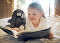 Happy smiling little girl lying on a bed at home and reading a childrens book to her toy teddy. Cute child relaxing and