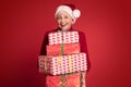 mature woman in red santa claus hat holding stack of gift boxes, on red studio background. Christmas shopping, copy Royalty Free Stock Photo