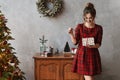 Happy smiling model girl in plaid red dress holding the present box and posing near Christmas tree. Young beautiful Royalty Free Stock Photo