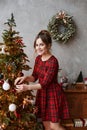 Happy smiling model girl with trendy hairstyle in plaid red dress decorating Christmas tree. Young beautiful woman in Royalty Free Stock Photo