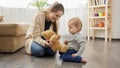 Happy smiling mother playing with her baby son with teddy bear on floor in living room. Royalty Free Stock Photo