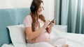 Happy smiling pregnant woman listening to music with headphones and using smartphone while sitting on bed at home Royalty Free Stock Photo