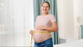 Happy smiling pregnant woman standing at window in bedroom and measuring big belly with measuring tape Royalty Free Stock Photo