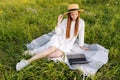 Happy smiling red-haired young woman wearing straw hat and white dress looking to laptop screen sitting on beautiful Royalty Free Stock Photo