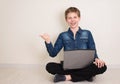 Happy smiling teenager boy working on laptop computer while sitting on the floor with legs crossed and pointing finger away Royalty Free Stock Photo