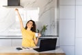 Happy smiling woman sitting at table in kitchen drinking morning coffee reading message looking at device screen using computer Royalty Free Stock Photo