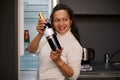 Happy smiling woman standing by refrigerator, holding a bottle of wine with white blank mockup label, choosing drink for Royalty Free Stock Photo