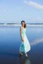 Happy smiling woman walking barefoot on empty beach. Full body portrait. Slim Caucasian woman wearing long dress. Blue sky. Summer