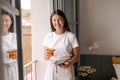 Happy smiling young caucasian woman wears pajamas holding smartphone and orange fresh at home. Royalty Free Stock Photo