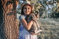 Happy smiling young girl in white hat and dress with flowers print holding a white and grey cat on her hands, looking at Royalty Free Stock Photo