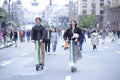 Happy smiling young man and girl ride electric scooters on the street, blurred people walking on a background