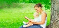 Happy smiling young mother and baby son reading book together on the grass under tree in summer park Royalty Free Stock Photo