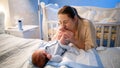 Happy smiling young mother looking at her newborn baby boy lying on bed at night and kissing his feet. Concept of family Royalty Free Stock Photo