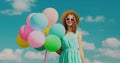 Happy smiling young woman with bunch of colorful balloons wearing a summer straw hat on the field on blue sky background Royalty Free Stock Photo