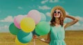 Happy smiling young woman with bunch of colorful balloons wearing a summer straw hat on the field on blue sky background Royalty Free Stock Photo