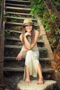 Happy smiling young woman dressed in hat and white long dress sitting barefoot on a vintage wooden stairs. Royalty Free Stock Photo