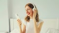 Happy smiling young woman listening to music with wireless headphones looking at phone on bed in white room at home Royalty Free Stock Photo