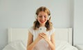 Happy smiling young woman listening to music with wireless headphones looking at smartphone on bed in white room at home Royalty Free Stock Photo