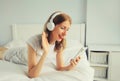 Happy smiling young woman listening to music with wireless headphones looking at smartphone on bed in white room at home Royalty Free Stock Photo