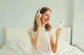 Happy smiling young woman listening to music with wireless headphones looking at smartphone on bed in white room at home Royalty Free Stock Photo