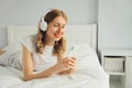 Happy smiling young woman listening to music with wireless headphones looking at smartphone on bed in white room at home Royalty Free Stock Photo