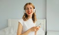 Happy smiling young woman listening to music with wireless headphones looking at smartphone on bed in white room at home Royalty Free Stock Photo