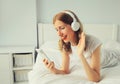 Happy smiling young woman listening to music with wireless headphones looking at smartphone talking on video call on bed in white Royalty Free Stock Photo