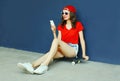 Happy smiling young woman with phone sitting on skateboard wearing red baseball cap, shorts on city street over blue wall Royalty Free Stock Photo