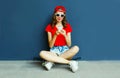 Happy smiling young woman with phone sitting on skateboard wearing red baseball cap, shorts on city street over blue wall Royalty Free Stock Photo