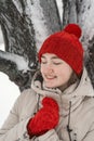 Happy smiling young woman in red hat and mittens stands near snow-covered tree. Portrait of cute thoughtfully girl with closed