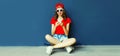 Happy smiling young woman with smartphone sitting on skateboard wearing red baseball cap, shorts on city street on blue wall Royalty Free Stock Photo