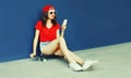 Happy smiling young woman with smartphone sitting on skateboard wearing red baseball cap, shorts on city street on blue wall Royalty Free Stock Photo