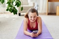 Happy smiling young woman in sportwear relaxing on mat on floor and using mobile cellphone at home, copy space. Full length body. Royalty Free Stock Photo