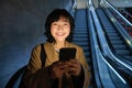 Happy smiling young woman, standing on escalator, going down, holding smartphone in both hands, chatting on mobile phone Royalty Free Stock Photo
