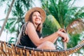 Happy smiling young woman in straw hat sitting in hammock with palm trees background Royalty Free Stock Photo