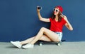Happy smiling young woman taking selfie picture by smartphone sitting on skateboard wearing red baseball cap, shorts on city Royalty Free Stock Photo