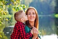 Happy son on the background of the lake hugs a young smiling mother behind the neck and kisses on the cheek. Parenting, family day Royalty Free Stock Photo