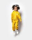 Happy sporty dark-skinned curly mulatto kid girl in warm comfortable yellow sport jumpsuit does fitness exercises jumps