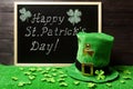 Happy St. Patricks Day chalk lettering on blackboard, green leprechaun hat and shamrock leaves on green grass. Dark wooden planks Royalty Free Stock Photo