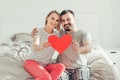 Happy strong marriage. Happy funny couple in love holding red paper heart. Heterosexual family man and woman sitting on bed in Royalty Free Stock Photo