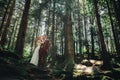 Happy stylish couple newlyweds in the green forest on summer day. bride in long white dress and groom in red suit are hugging.