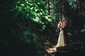 Happy stylish couple newlyweds in the green forest on summer day. bride in long white dress and groom in red suit are hugging.