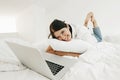 Happy stylish girl lying on white bed with laptop and smiling. Joyful young woman hugging pillow and looking at laptop. Watching Royalty Free Stock Photo