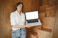 Happy stylish girl showing laptop with empty screen, standing at wooden stairs in modern room. Young business woman working online Royalty Free Stock Photo