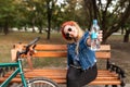 Happy, stylish girl sitting on a bench in a park near a bicycle and shows a bottle of water in the camera Royalty Free Stock Photo